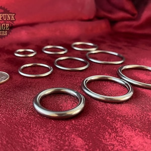 May include: A set of eight silver metal rings of varying sizes, displayed on a red fabric surface. The rings are smooth and round, with a polished finish. A US quarter is included for scale.