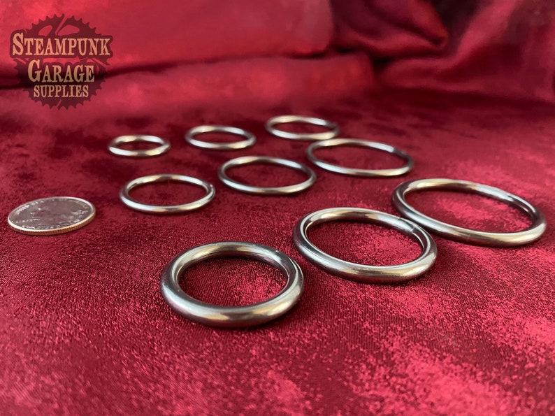May include: A set of eight silver metal rings of varying sizes, displayed on a red fabric surface. The rings are smooth and round, with a polished finish. A US quarter is included for scale.