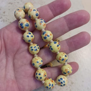 Puede incluir: Una pulsera de cuentas crema con dise&ntilde;os florales azules y naranjas en cuentas redondas. Las cuentas est&aacute;n ensartadas en un cord&oacute;n beige. La pulsera se sostiene en una mano.