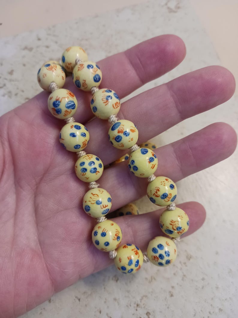 Puede incluir: Una pulsera de cuentas crema con dise&ntilde;os florales azules y naranjas en cuentas redondas. Las cuentas est&aacute;n ensartadas en un cord&oacute;n beige. La pulsera se sostiene en una mano.
