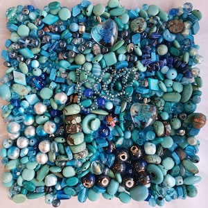 May include: A large collection of loose beads in various shades of blue, turquoise, and teal. The assortment includes round, oval, and irregular shapes, featuring two clear heart-shaped pendants with blue floral inclusions and a beaded chain.