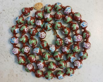 Puede incluir: Un collar con cuentas redondas esmaltadas en rojo, verde y blanco, decoradas con motivos florales. Las cuentas están ensartadas en un cordón marrón rojizo y se cierran con un cierre dorado. El collar es de estilo vintage.