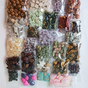 May include: A collection of assorted loose beads in clear plastic bags, featuring various shapes, sizes, and materials including wood, stone, shell, and glass in colors like brown, green, pink, blue, white, and black.