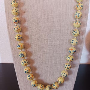 Puede incluir: Un collar de cuentas con grandes cuentas redondas de color amarillo p&aacute;lido. Cada cuenta est&aacute; adornada con un patr&oacute;n de dise&ntilde;os florales y de puntos azules y naranjas. El collar se exhibe sobre una tela de color neutro.