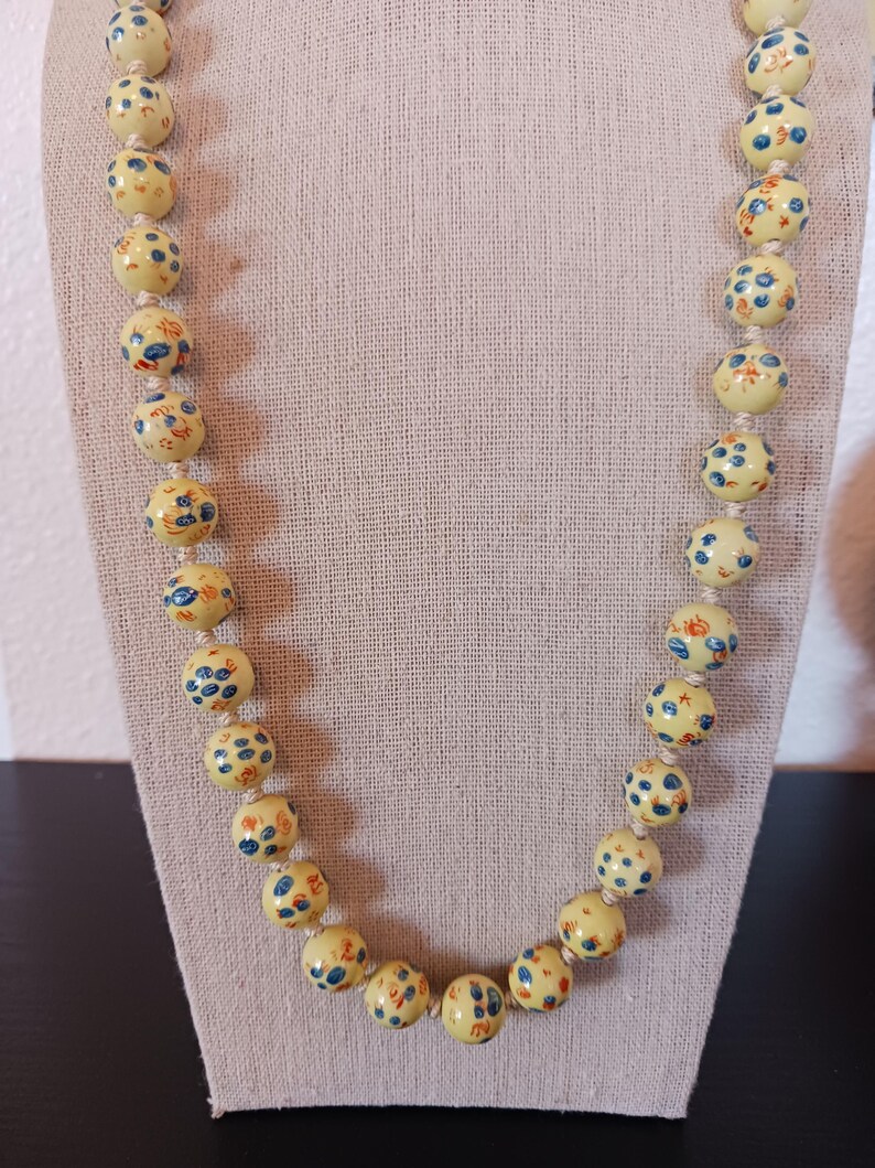 Puede incluir: Un collar de cuentas con grandes cuentas redondas de color amarillo p&aacute;lido. Cada cuenta est&aacute; adornada con un patr&oacute;n de dise&ntilde;os florales y de puntos azules y naranjas. El collar se exhibe sobre una tela de color neutro.