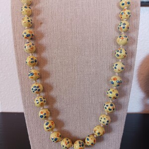 Puede incluir: Un collar largo con cuentas redondas de color amarillo p&aacute;lido ensartadas en un cord&oacute;n claro. Cada cuenta est&aacute; decorada con peque&ntilde;os motivos florales o abstractos pintados a mano en azul y naranja, distribuidos uniformemente.