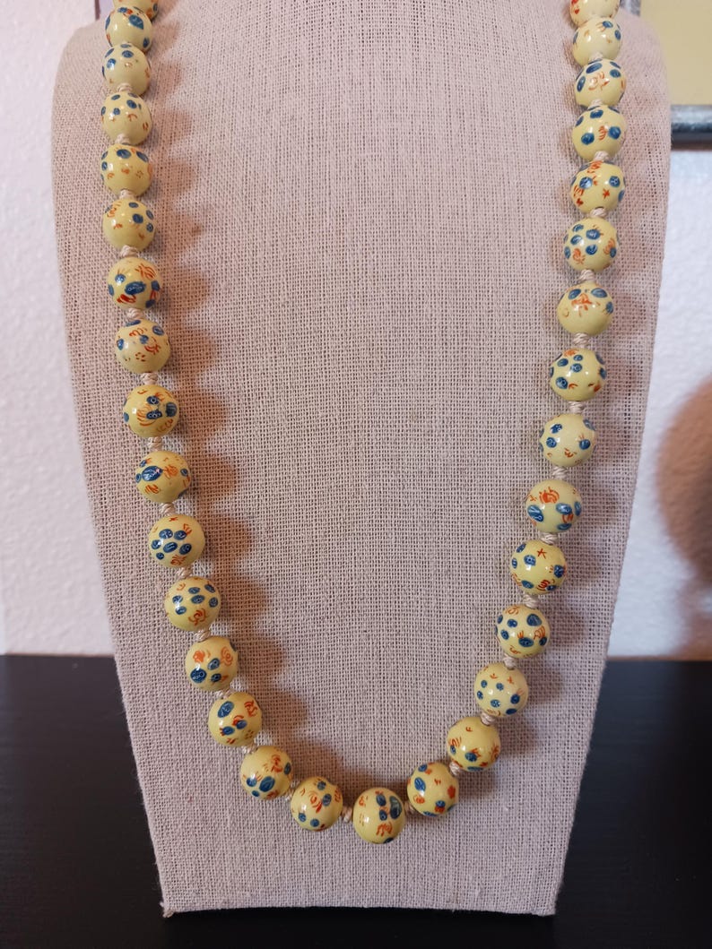 Puede incluir: Un collar largo con cuentas redondas de color amarillo p&aacute;lido ensartadas en un cord&oacute;n claro. Cada cuenta est&aacute; decorada con peque&ntilde;os motivos florales o abstractos pintados a mano en azul y naranja, distribuidos uniformemente.