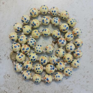 Puede incluir: Un collar con cuentas redondas de color crema decoradas con peque&ntilde;os motivos florales pintados a mano en azul y naranja. Las cuentas est&aacute;n ensartadas en un cord&oacute;n claro e incluyen un peque&ntilde;o componente de cierre met&aacute;lico circular.