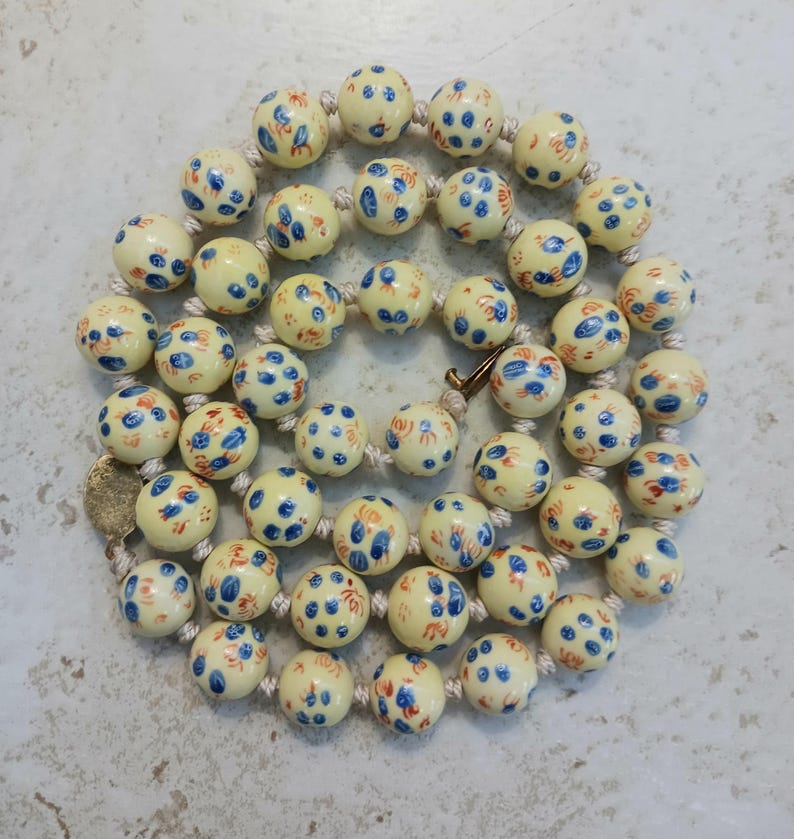 Puede incluir: Un collar con cuentas redondas de color crema decoradas con peque&ntilde;os motivos florales pintados a mano en azul y naranja. Las cuentas est&aacute;n ensartadas en un cord&oacute;n claro e incluyen un peque&ntilde;o componente de cierre met&aacute;lico circular.