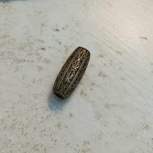 May include: A bronze-colored, elongated bead with intricate filigree patterns. The bead has a cylindrical shape with detailed openwork designs, featuring floral and geometric motifs. The metal bead is likely used for jewelry making or decorative purposes.