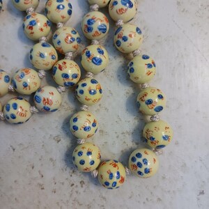 Puede incluir: Un collar con cuentas redondas de color crema decoradas con patrones florales azules y rojos. Las cuentas est&aacute;n ensartadas en un cord&oacute;n blanco. El collar es de estilo vintage.