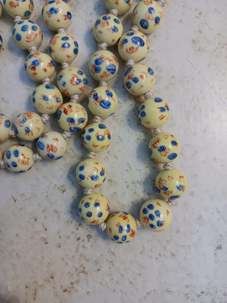 Puede incluir: Un collar con cuentas redondas de color crema decoradas con patrones florales azules y rojos. Las cuentas est&aacute;n ensartadas en un cord&oacute;n blanco. El collar es de estilo vintage.