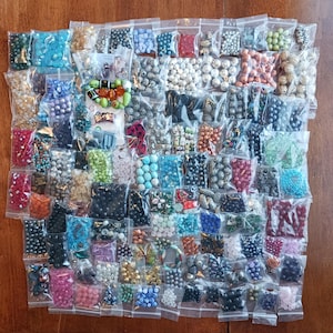 May include: A collection of various beads in small, clear plastic bags. The beads are in a variety of colors, including blue, green, red, and silver. Some bags contain multiple bead shapes and sizes. The beads are arranged on a brown wooden surface.