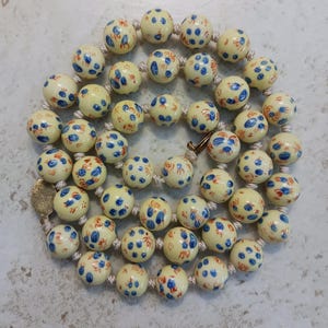 Puede incluir: Un collar con cuentas redondas de color crema decoradas con peque&ntilde;os motivos florales pintados a mano en azul y naranja. Las cuentas est&aacute;n ensartadas en un cord&oacute;n claro, con un cierre de metal dorado y una peque&ntilde;a etiqueta met&aacute;lica redonda.