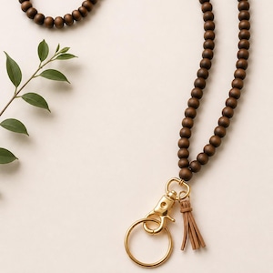 May include: A lanyard featuring a long strand of round, dark brown wooden beads. At the base, a gold-toned metal lobster clasp connects to a circular gold-toned key ring and a small, light tan faux leather tassel, creating a functional accessory.