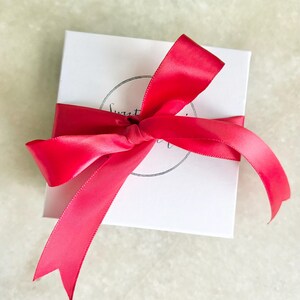May include: A white gift box with a bright pink satin ribbon tied in a bow. The box has a black circle logo with the text "Sweet" and "Treat" inside.