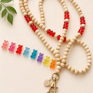 May include: A lanyard featuring natural light-colored wooden beads interspersed with translucent red gummy bear-shaped charms. The lanyard includes a gold-toned metal lobster clasp with a small circular charm engraved with the letter R.