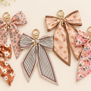 May include: Five fabric bow keychains featuring various patterns including floral prints in pink, orange, and white, and a black and white striped design. Each bow is attached to a gold-toned metal ring and accented with small white faux pearl beads.