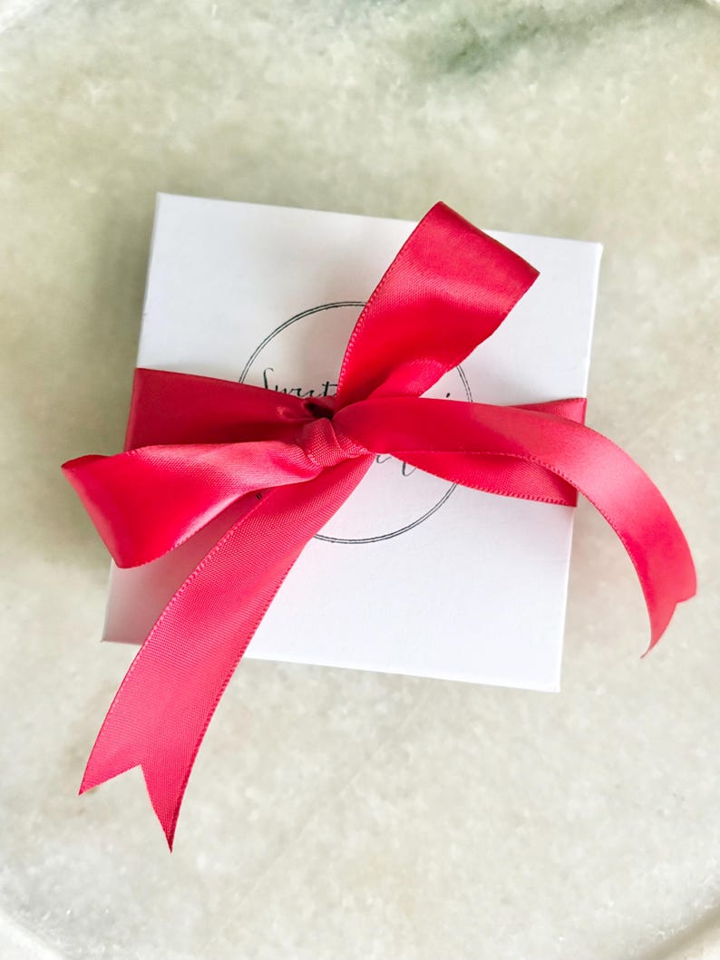 May include: A white gift box with a bright pink satin ribbon tied in a bow. The box has a black circle logo with the text "Sweet" and "Treat" inside.