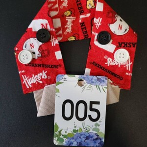 May include: Red and white fabric headband with the words "Nebraska Huskers" and a football helmet design. The headband has white buttons and a white tag with the number "005" printed on it.