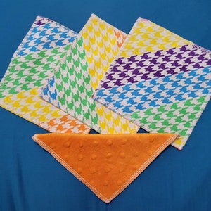 May include: A collection of colorful, square fabric cloths with a houndstooth pattern in rainbow hues. An orange, triangular cloth with a textured surface is also present. The cloths are arranged on a blue surface.