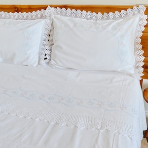 May include: White bedding set with delicate crochet trim and embroidered floral details. The set includes a flat sheet, fitted sheet, and two pillowcases.