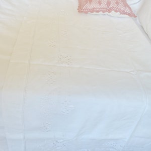 May include: White cotton bed sheet with delicate embroidered floral designs along the edge. The sheet is folded and a pink crocheted pillow is on top.