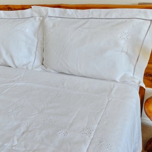 May include: White embroidered bedding with two white pillowcases on a wooden bed frame. A vase with white flowers sits on a wooden side table.