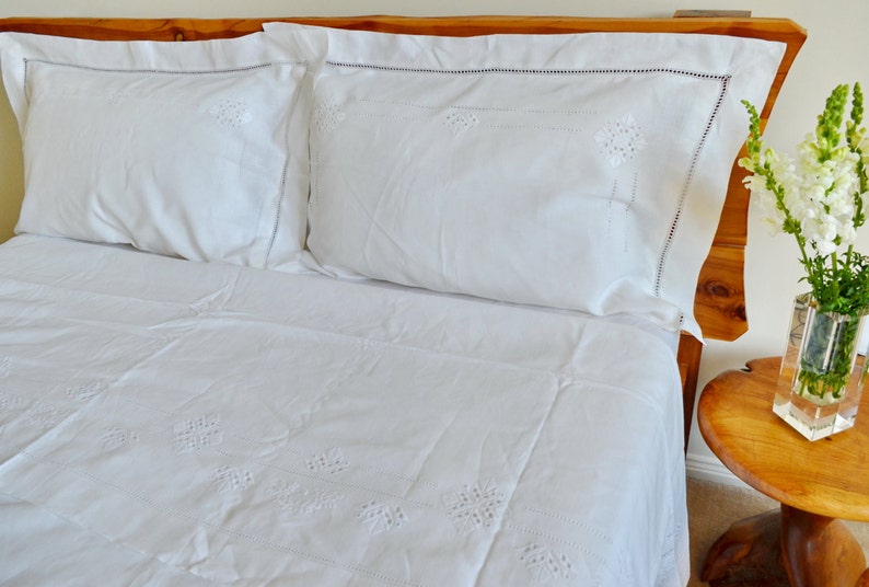 May include: White embroidered bedding with two white pillowcases on a wooden bed frame. A vase with white flowers sits on a wooden side table.