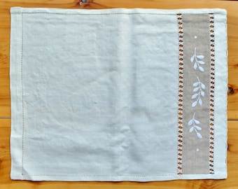 Puede incluir: Un mantel individual de lino blanco con un borde beige y hojas blancas bordadas. El mantel individual tiene un diseño simple y elegante.
