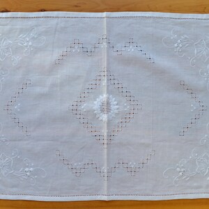Könnte beinhalten: Weiße bestickte Tischdecke aus Leinen mit floralem Muster und einem zentralen Diamantenmuster. Die Tischdecke hat ein zartes, filigranes Design und ist perfekt, um jeder Tischdekoration einen Hauch von Eleganz zu verleihen.