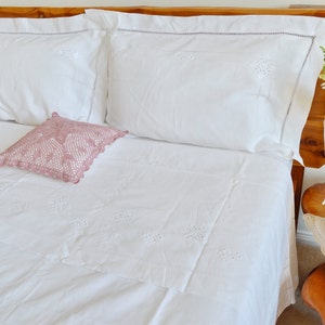 May include: White bedding with delicate embroidered details, featuring a pink crocheted pillow and a wooden headboard.