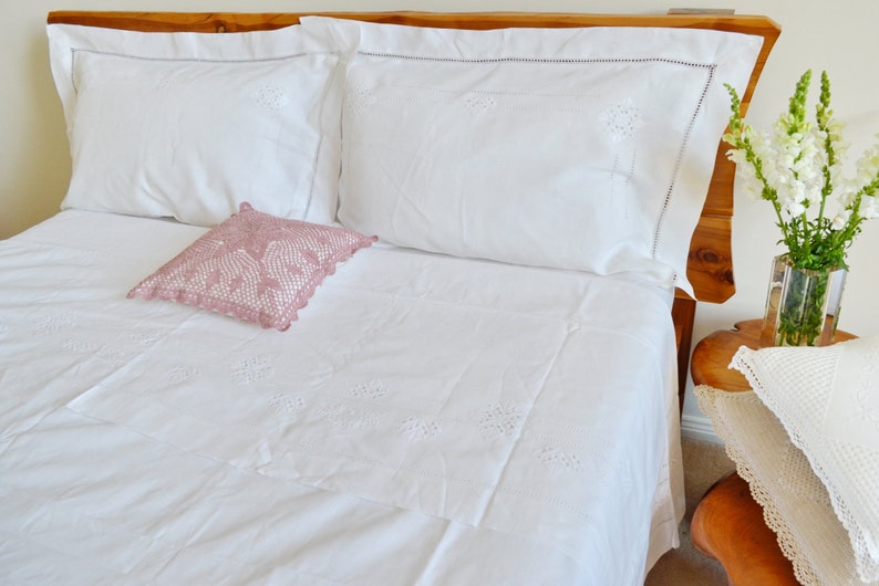May include: White bedding with delicate embroidered details, featuring a pink crocheted pillow and a wooden headboard.