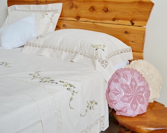 May include: A white bed with a wooden headboard and embroidered bedding. The bed is made with a white sheet and a white pillow with lace trim. There are two crocheted pillows on a wooden stool next to the bed.