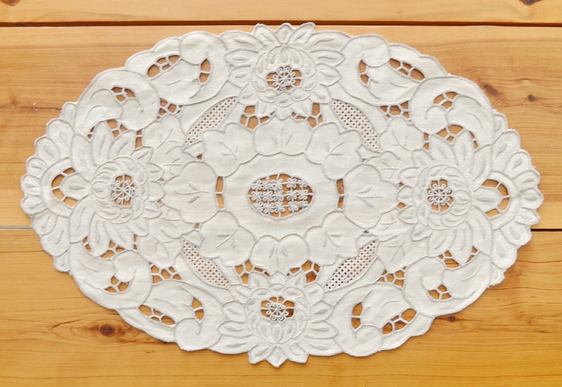 Puede incluir: Mantel de encaje blanco ovalado con un delicado dise&ntilde;o floral. El encaje es intrincado y presenta un patr&oacute;n repetitivo de flores y hojas.