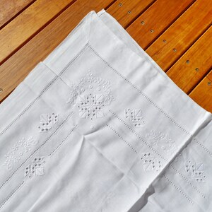 May include: White linen tablecloth with delicate embroidered floral designs. The tablecloth is folded in half and has a subtle sheen.