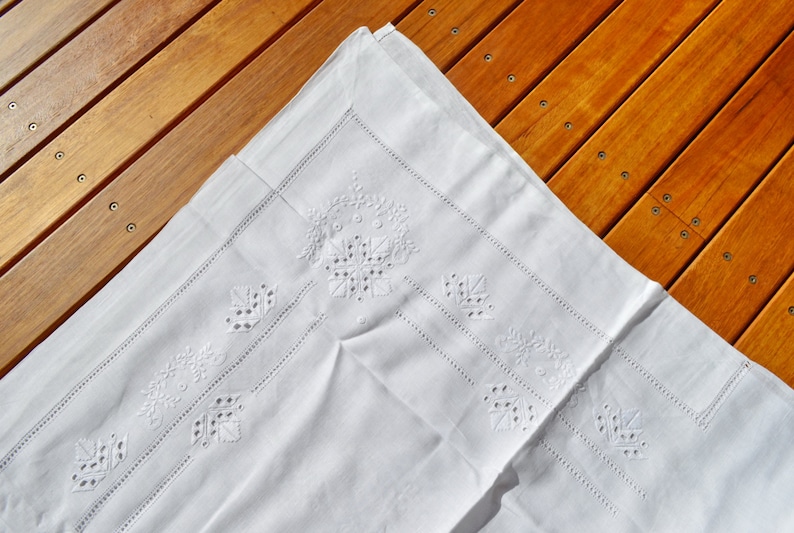 May include: White linen tablecloth with delicate embroidered floral designs. The tablecloth is folded in half and has a subtle sheen.