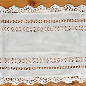 以下が含まれることがあります： 繊細なレースの縁取りが施された、白いクロシェのテーブルランナー。ランナーは長方形で、薄くて軽い生地で作られています。