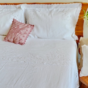 May include: A white bed with a white crocheted bedspread and two white pillows with lace trim. A pink crocheted pillow is on top of the bed. There is a wooden headboard and a small wooden table with a white crocheted pillow and a vase of white flowers.