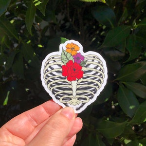 May include: A sticker featuring a heart-shaped rib cage design with colorful flowers. The flowers include red, orange, and purple blooms with green leaves. The sticker is held in a hand against a background of green leaves.