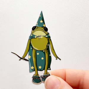 Pu&ograve; includere: Una rana verde che indossa un cappello da mago e un mantello con stelle blu e gialle, tenendo un bastone. La rana indossa scarpe blu con lacci gialli.