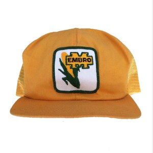 May include: A yellow trucker hat with a green and white embroidered patch that says "EMBRO" and depicts a corn stalk. The hat has a mesh back.