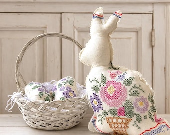 May include: A white fabric Easter bunny with floral cross-stitch embroidery, next to a gray wicker basket filled with decorated eggs. The bunny has red and blue accents on its ears. The eggs and bunny feature purple, pink, and green floral designs.