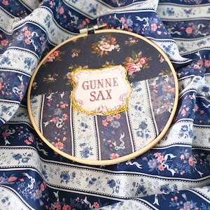 May include: A round embroidery hoop with a floral fabric design. The fabric is a patchwork of different floral prints in shades of blue, pink, and white. The center of the hoop features a rectangular patch with the text "GUNNE SAX" embroidered in red thread.