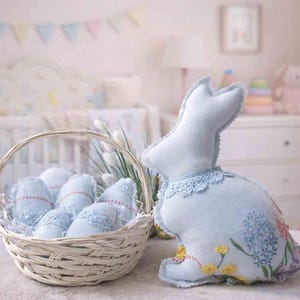 Pode incluir: Um coelho de pelúcia azul claro da Páscoa com detalhes florais bordados e uma gola de renda está ao lado de uma cesta de vime cheia de ovos de Páscoa de tecido azul. A cena é ambientada em um quarto de bebê em tons pastel.