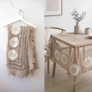 May include: A beige crochet tablecloth featuring intricate openwork patterns and circular fabric inserts embroidered with floral motifs. The tablecloth is displayed hanging on a white hanger and draped over a wooden table in a bright room.