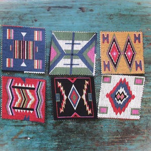 Puede incluir: Seis posavasos cuadrados de tela con patrones geométricos en varios colores. Los diseños incluyen diamantes, zigzags y rayas en tonos de azul, rojo, negro, verde y dorado. Los posavasos están dispuestos sobre una superficie turquesa desgastada.