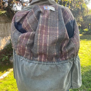 K&ouml;nnte beinhalten: Gr&uuml;ne Carhartt-Jacke mit kariertem Innenfutter. Die Jacke hat einen Kragen und eine Taille mit Kordelzug. Das Innere ist eine Mischung aus roten, braunen und grauen Karos. Die Jacke ist drau&szlig;en an einem sonnigen Tag.