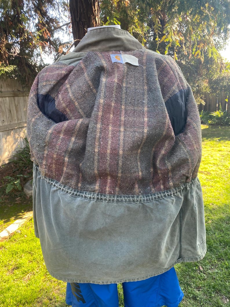 K&ouml;nnte beinhalten: Gr&uuml;ne Carhartt-Jacke mit kariertem Innenfutter. Die Jacke hat einen Kragen und eine Taille mit Kordelzug. Das Innere ist eine Mischung aus roten, braunen und grauen Karos. Die Jacke ist drau&szlig;en an einem sonnigen Tag.