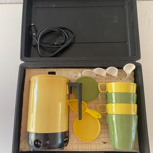 May include: A black plastic carrying case containing a yellow and black electric kettle, four stacked plastic cups in alternating yellow and green colors, four white spoons, a yellow scoop, two circular lids in green and yellow, and a power cord.